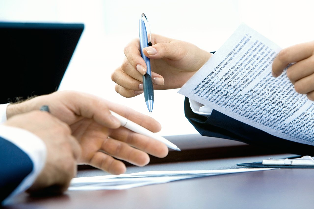 Two people at a desk, one holding a pen over a printed document and the other handing across a second pen.
