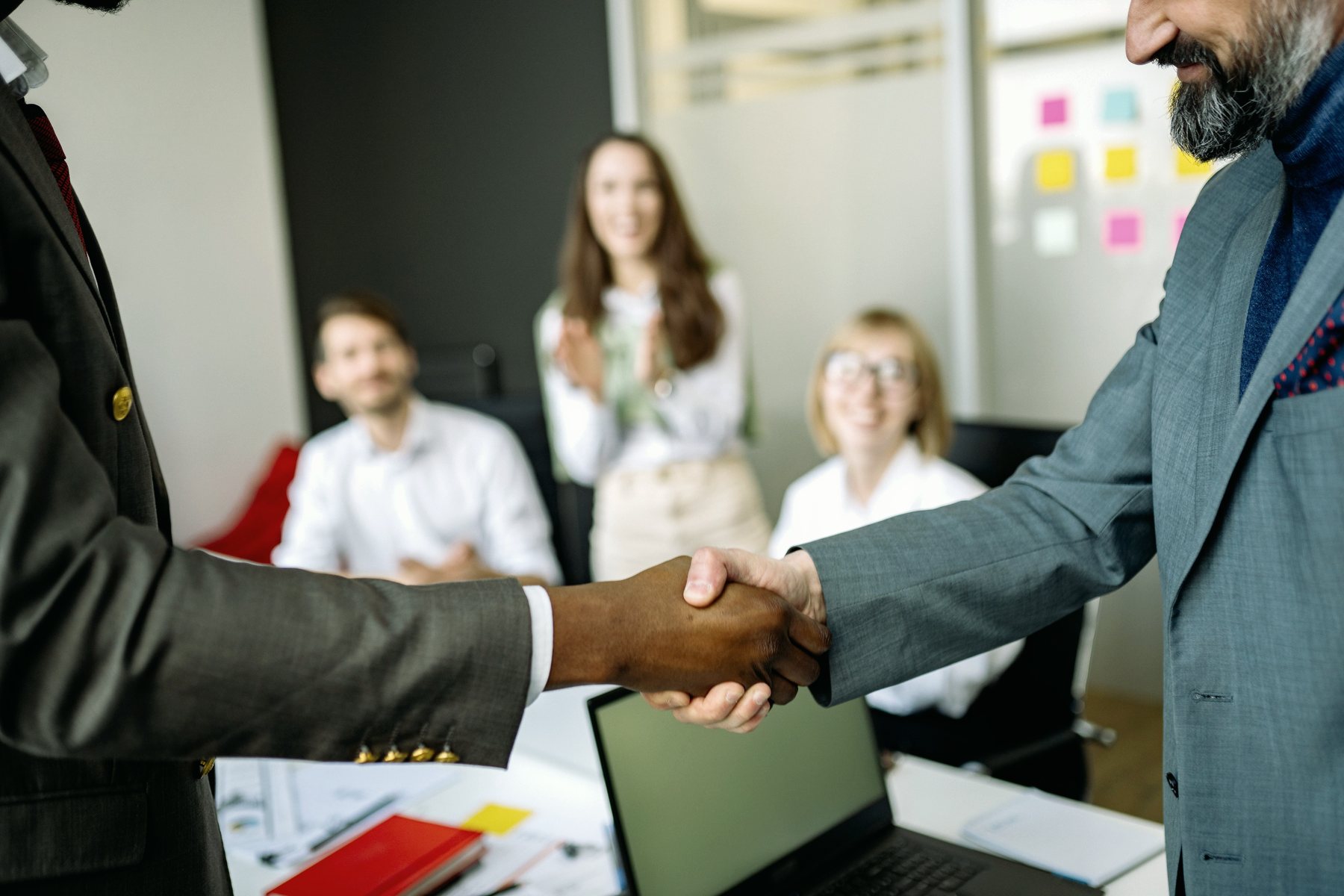 Two people in suits shaking hands across a desk while colleagues applaud in the background.