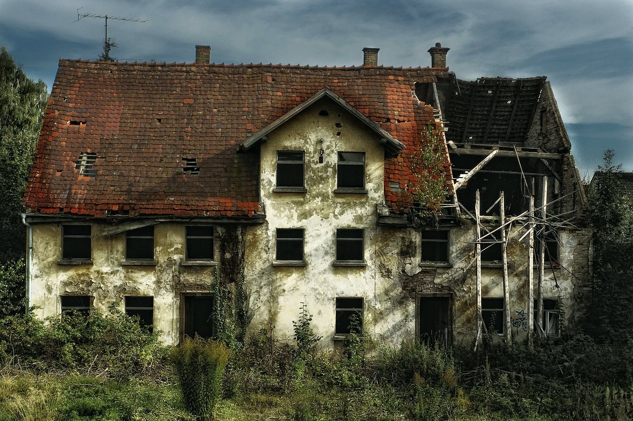 A derelict house with a collapsed roof and empty window frames, overgrown with weeds.