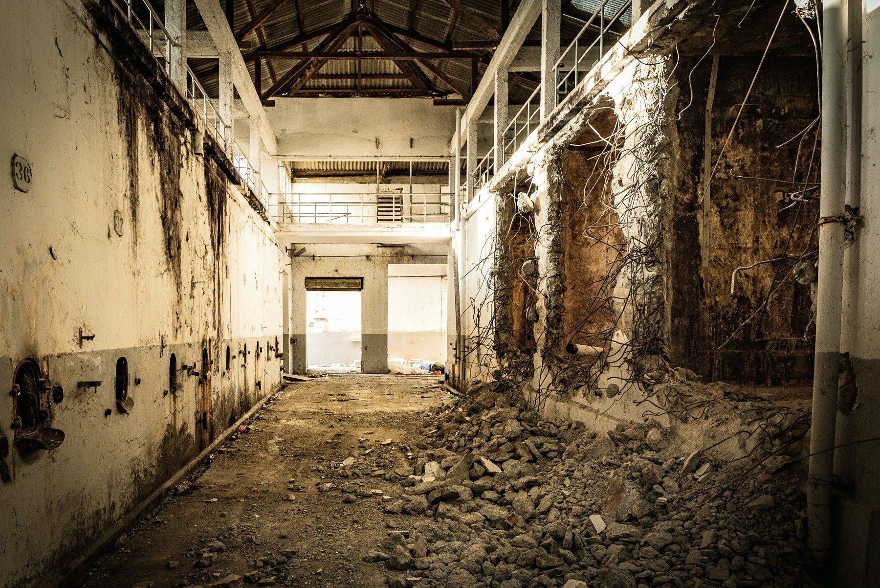The inside of an abandoned factory, with rubble on the floor and wires hanging from a broken wall.