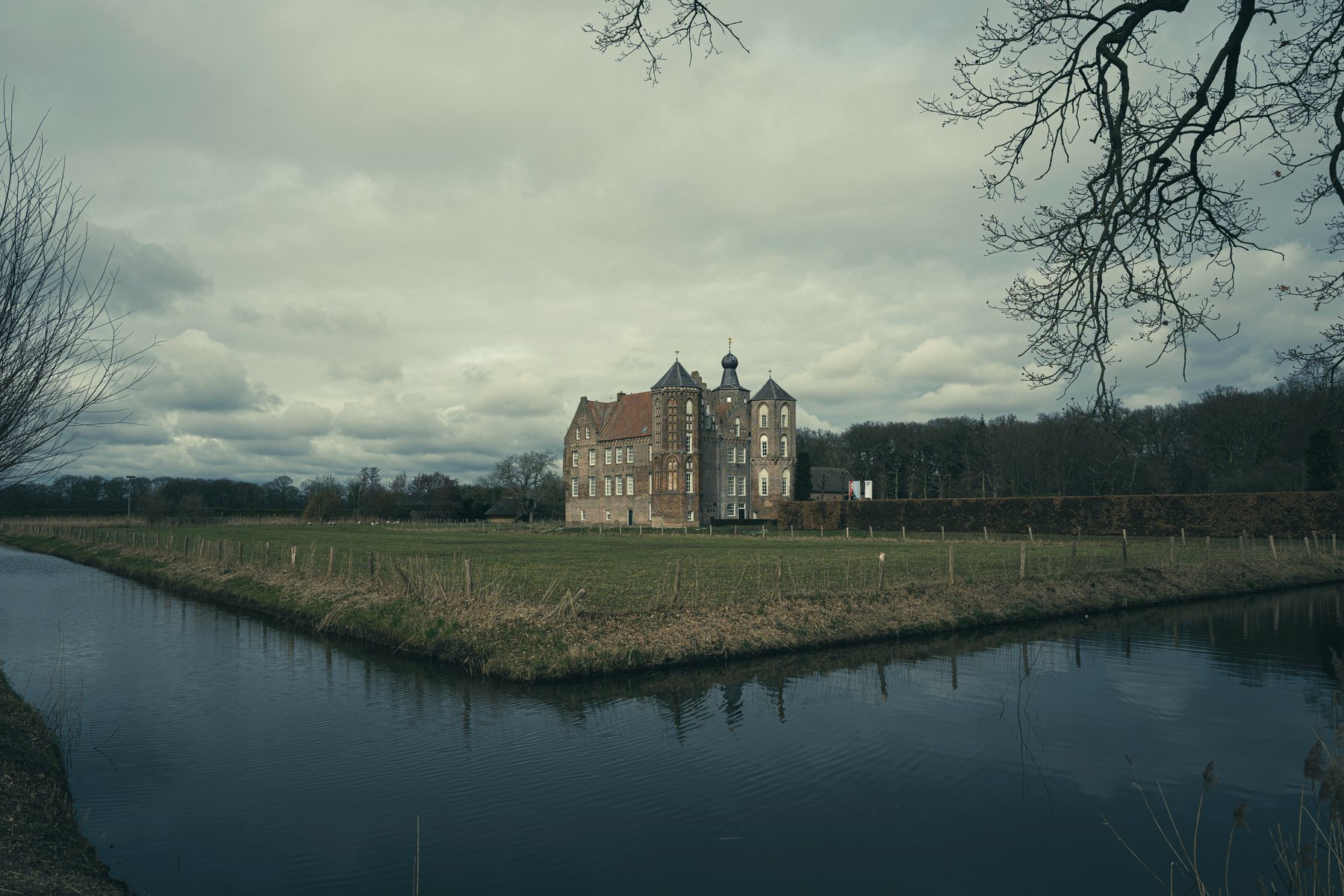 A brick castle with turrets standing in a field, surrounded by a still moat under a grey sky.