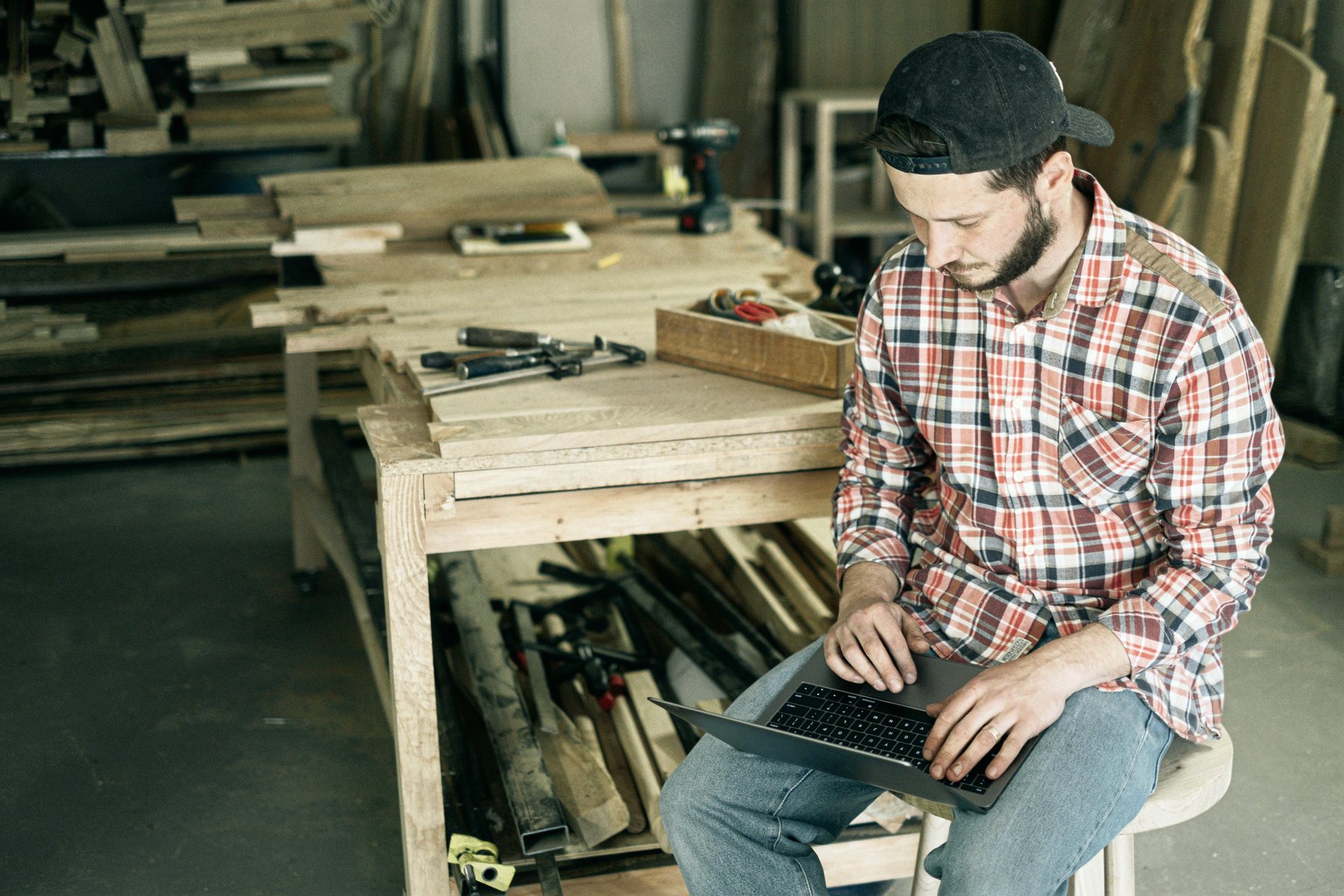 A carpenter working on a laptop at a bench in their own workshop.