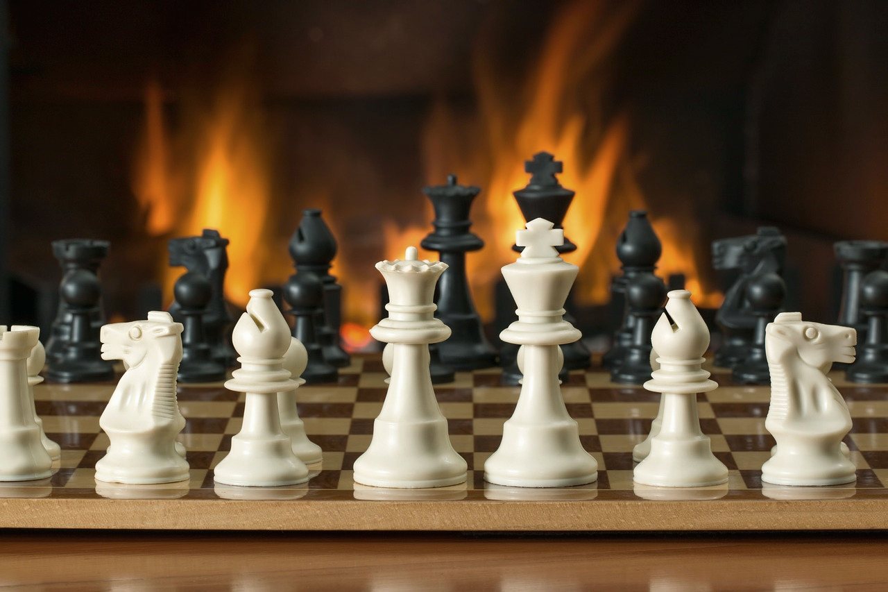 Chess pieces lined up on a wooden board in front of a fireplace.